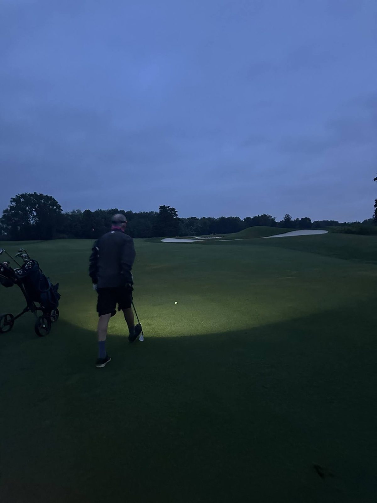 A golfer walking a dark fairway lit by a headlamp